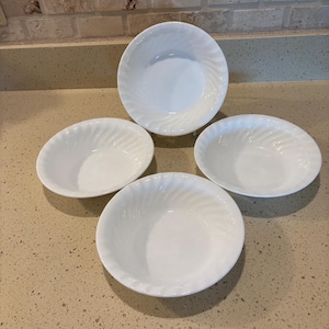 May include: A set of four white bowls featuring a subtle, swirling spiral pattern embossed along the interior rim. Each bowl has a smooth, glossy finish and a rounded shape, arranged in a cluster to display the uniform design and bright white color.