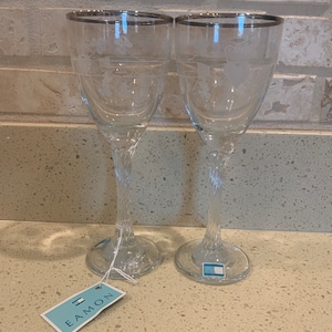 May include: Two clear glass wine glasses with a silver rim and a twisted stem. The glasses have an etched design and are sitting on a light brown countertop. A blue and white tag with the word "Eamon" is in the foreground.