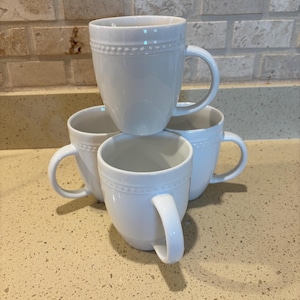 May include: Four white ceramic mugs stacked together. Each mug has a rounded body, a curved handle, and a decorative beaded band near the rim. The mugs are arranged on a light-colored countertop with a neutral-toned brick backsplash.