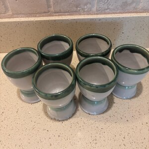 May include: Six ceramic egg cups. Each cup is white with a green rim and base. The cups are arranged in a group on a speckled countertop.