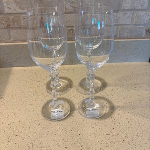 May include: Two clear crystal wine glasses with a decorative stem and a round base. Each glass has a smaller glass inside. The base of each glass has a white label with the text "VERA WANG" and "WEDGWOOD".