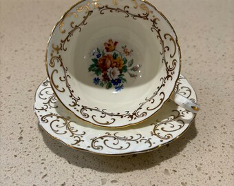 May include: A white teacup and saucer with a gold floral design. The cup and saucer have a scalloped edge and a delicate floral pattern in gold.