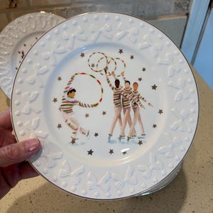 May include: A white ceramic plate with a silver rim and a raised bow and dot pattern along the border. The center features a colorful illustration of four figures in striped sweaters and light-colored pants performing with hula hoops among gold stars.