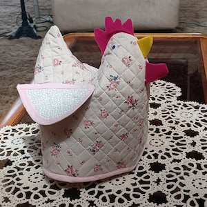 May include: A quilted chicken-shaped pot holder with a pink and yellow beak, pink feet, and a floral pattern. The pot holder is sitting on a white crocheted doily.