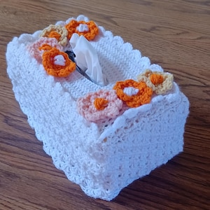 May include: White crocheted tissue box cover with a crocheted flower design in orange, yellow, and pink.