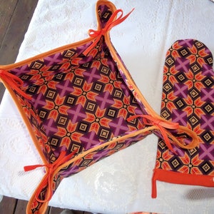 May include: A set of two orange and purple patterned kitchen accessories. The first item is a square-shaped bread basket with orange fabric handles. The second item is a matching oven mitt.