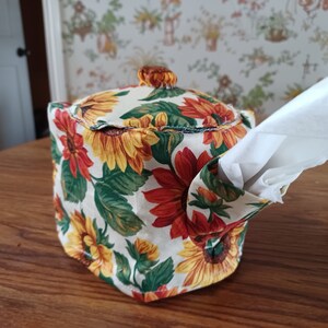 May include: A white fabric tissue box cover with a sunflower pattern. The cover is shaped like a teapot and has a cutout for tissues.