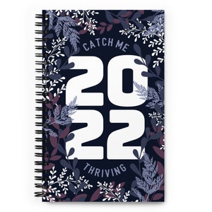 May include: A spiral-bound notebook with a dark blue cover featuring the text "CATCH ME 2022 THRIVING" in white. The text is surrounded by a floral design in white, purple, and blue. The notebook is designed for planning and journaling.