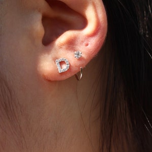Puede incluir: Primer plano de la oreja de una persona con tres pendientes. Los pendientes son un pequeño pendiente de diamante, un pendiente de aro y un pendiente con la letra D con pequeños diamantes.