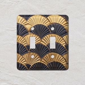 May include: A double light switch cover featuring a repeating fan-shaped design in gold and black. The cover has two switch openings and screw holes. The design is reminiscent of the Art Deco style.