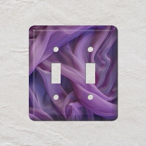 May include: A decorative double light switch cover featuring a flowing, abstract design in shades of purple and lavender. The cover has two toggle switch openings and four screw holes. The background is a textured white surface.