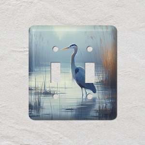 May include: A light switch cover with a wetland design. A blue heron stands in the water, with reeds and grasses in the background. The cover has two switch openings and four screw holes. The finish is glossy.