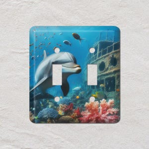 May include: A decorative light switch cover featuring a vibrant underwater scene. A smiling dolphin swims near a coral reef and a sunken ship. The cover has two switch openings and is painted in shades of blue, green, and orange.