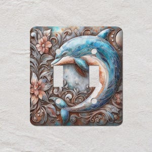 May include: A decorative light switch cover featuring a blue dolphin design. The dolphin is surrounded by floral and scrollwork elements in shades of brown, blue, and peach. The cover has two switch openings.