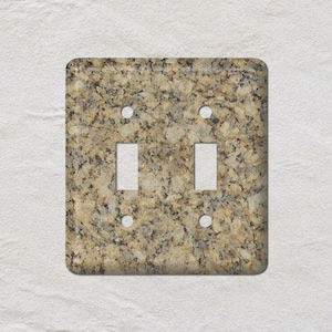 May include: A square, double-toggle light switch cover with a granite-like pattern in shades of tan, brown, and black. The cover has two rectangular openings for the switches and four small circular holes for mounting.