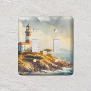 May include: A decorative light switch cover featuring a watercolor painting of a lighthouse on a rocky coastline. The lighthouse has a red and white striped tower, with a small building at its base. The scene includes trees, the ocean, and a cloudy sky.