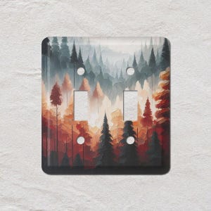 May include: A square light switch cover featuring a painted forest scene. The artwork depicts tall evergreen trees in shades of red, orange, and gray. The cover has two switch openings and two screw holes.