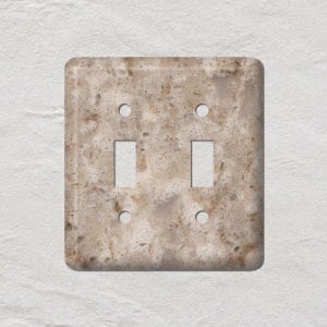 May include: A double light switch cover with a beige and brown speckled pattern, resembling natural stone. The cover has two rectangular switch openings and four screw holes. The edges are rounded.