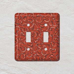 May include: A decorative double light switch cover with a retro boomerang pattern. The cover is a vibrant red with a repeating design of black and cream-colored boomerang shapes. The cover has two switch openings and four screw holes.