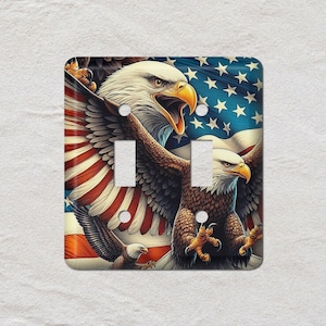 May include: A decorative light switch cover featuring a patriotic design. The cover showcases two bald eagles with outstretched wings against a backdrop of the American flag. The eagles are depicted with detailed feathers and sharp beaks, with the flag's stars and stripes visible.