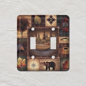 May include: A decorative double light switch cover with a rustic camping theme. The design features a collage of outdoor elements, including a bear, compass, lantern, and a canoe on a lake. The color palette includes browns, greens, and reds.