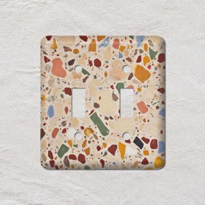 Peut inclure: Plaque d'interrupteur double carrée avec un motif terrazzo. La plaque présente une base crème avec des éclats colorés de rouge, orange, vert, bleu et jaune. Elle comporte deux découpes rectangulaires pour les interrupteurs et deux trous de vis.