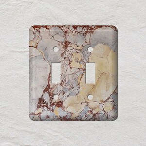 May include: A decorative double light switch cover with a marble design. The cover features a square shape with two rectangular cutouts for the switches and two small circular holes for the screws. The marble pattern includes shades of gray, brown, and yellow.