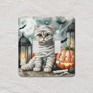 May include: Halloween-themed light switch cover featuring a cat dressed as a mummy. The design includes a jack-o'-lantern, bats, candles, and a full moon. The artwork is a watercolor style with a spooky, yet cute, aesthetic.