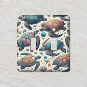 May include: A double light switch cover featuring a repeating pattern of colorful sea turtles, seashells, and starfish on a cream background. The turtles have blue, orange, and pink shells. The cover has two toggle switch openings.