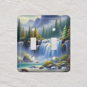 May include: A decorative double light switch cover featuring a scenic waterfall landscape. The artwork depicts a cascading waterfall with blue water, surrounded by green trees, rocks, and mountains. The cover has a glossy finish.