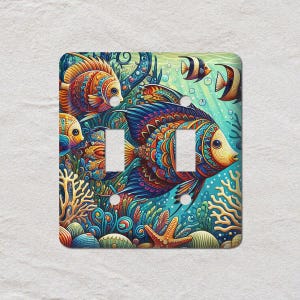 May include: A decorative double light switch cover with a colourful underwater design. Fish swim amongst coral and seaweed in shades of blue, orange, and yellow. The cover has two switch openings.
