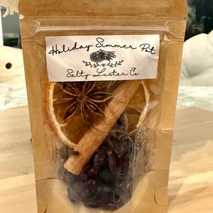 May include: A clear plastic bag filled with dried fruit and spices. The bag has a brown paper backing and a white label that reads "Holiday Simmer Pot" and "Salty Luster Co.". The contents include a dried orange slice, star anise, cinnamon stick, and dark berries.