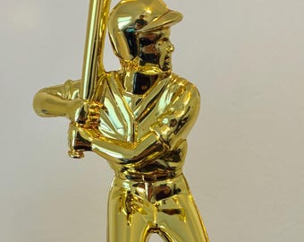 May include: A gold-colored baseball trophy featuring a baseball player in a batting stance, holding a bat. The trophy is mounted on a rectangular base.
