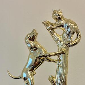 May include: A gold-colored trophy featuring a dog reaching up a tree towards a squirrel. The dog is on its hind legs, and the squirrel is perched on the tree. The trophy sits on a black and gold base.