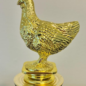 May include: A gold-colored chicken trophy. The chicken is perched atop a gold base, with a red and gold pedestal. The trophy is likely awarded for a poultry-related competition or event.