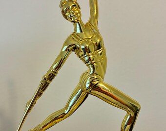 May include: A gold-colored trophy featuring a stylized figure in a dynamic pose, holding a staff and with one arm raised. The figure is mounted on a rectangular base, atop a multi-tiered pedestal.
