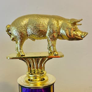 May include: A gold-colored trophy featuring a pig figurine. The pig is standing on a rectangular gold base, atop a decorative column with a purple and blue base. The pig has detailed fur texture.
