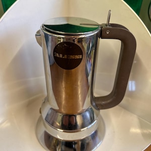 Puede incluir: Una cafetera espresso de acero inoxidable con asa y tapa marrones. La cafetera tiene una base redonda y un cuerpo cilíndrico. La marca "ALESSI" está impresa en una pegatina circular en la parte delantera.
