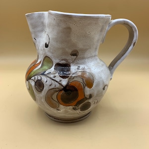 Könnte beinhalten: Eine weiße Keramikkanne mit einem braunen, orangefarbenen und grünen Blumendesign. Die Kanne hat einen leicht ausgestellten Rand und einen Henkel.