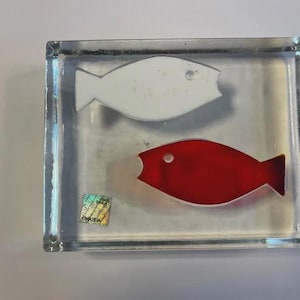 May include: A clear, rectangular block with two fish embedded inside. One fish is white, the other is red. The fish have simple, stylized shapes. The block has a slight shimmer and a holographic sticker is visible.