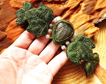 May include: A beaded green leaf bracelet featuring a large, carved, iridescent labradorite gemstone in the centre. The bracelet is on a hand, with the background out of focus.