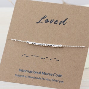 May include: Silver bracelet with the word "Loved" spelled out in International Morse Code using small beads. The bracelet is displayed on a brown card with the text "International Morse Code" and "Enjoyelry | Handmade for You | Silver 925".