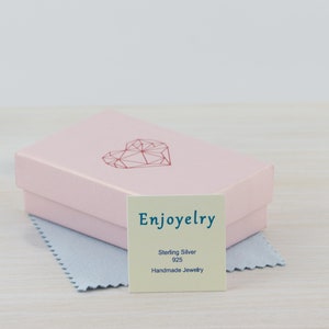 May include: A pink jewelry box with a geometric heart design on the lid. The box is open and a small white card with the text "Enjoyelry Sterling Silver 925 Handmade Jewelry" is visible.
