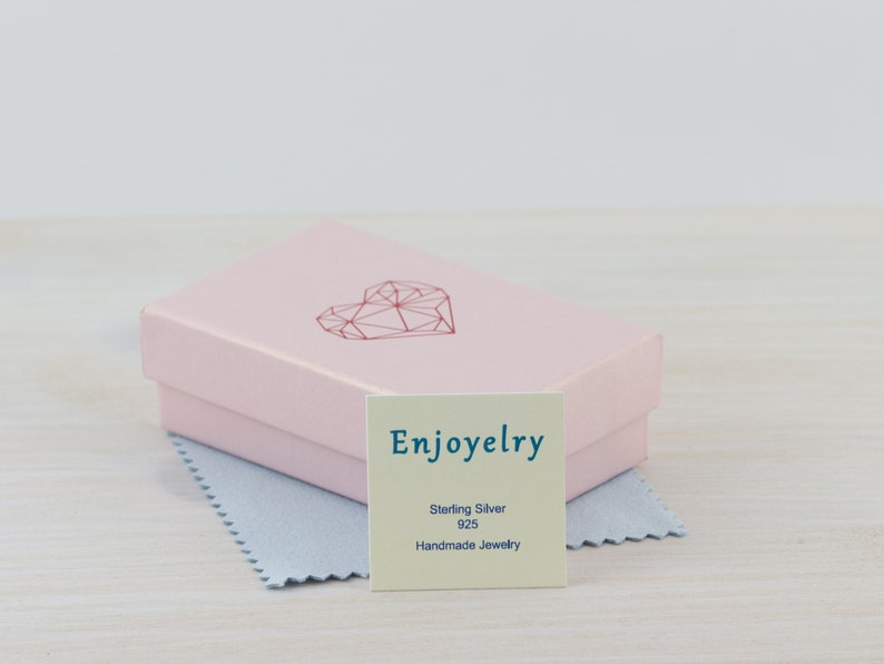 May include: A pink jewelry box with a geometric heart design on the lid. The box is open and a small white card with the text "Enjoyelry Sterling Silver 925 Handmade Jewelry" is visible.