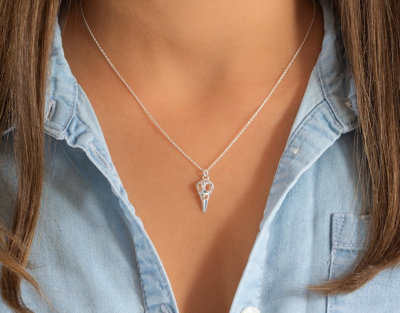 May include: A silver necklace with a delicate chain and a bird skull pendant. The pendant is detailed and intricate, with a hollow design.