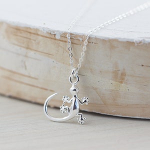 May include: A silver necklace with a detailed gecko pendant. The necklace is on a light wood surface.