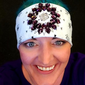 May include: White bandana with a paisley pattern, adorned with a central cluster of sparkling rhinestones and surrounding dark purple gems. The headwear is a stylish accessory.