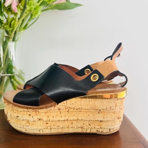 Puede incluir: Sandalias de plataforma de cuero negro con suela de corcho y ribete dorado. Las sandalias tienen un diseño de tiras cruzadas y una correa de tobillo de cuero marrón claro. La suela de plataforma mide unos 7.6 cm de alto.