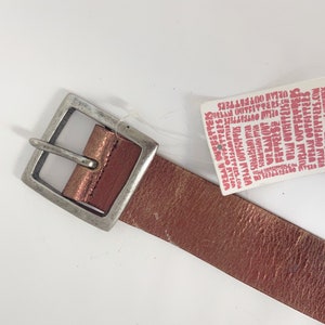 May include: A rose gold leather belt with a silver buckle. The belt has a tag attached that reads "Urban Outfitters".