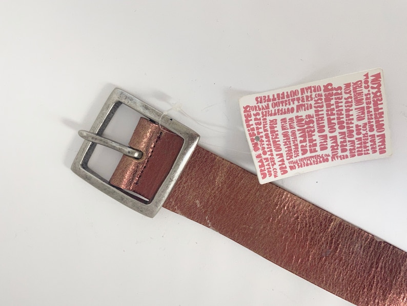 May include: A rose gold leather belt with a silver buckle. The belt has a tag attached that reads "Urban Outfitters".